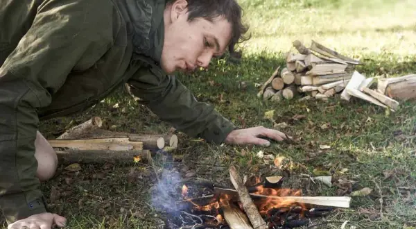 Mann entfacht mit einem Feuerstahl ein Lagerfeuer. Er pustet vorsichtig auf die Flammen, um sie zu nähren.