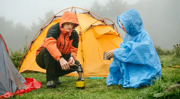 Zwei Personen im Regen bauen ein Zelt auf und bereiten sich ein warmes Getränk zu.