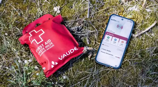 Rote Erste-Hilfe-Tasche und Smartphone mit Notruf-App liegen im Gras. Wichtige erste hilfe basics für unterwegs.