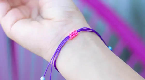 Eine Hand trägt ein lila Armband mit einem doppelten Schiebeknoten. Kleine blaue Perlen zieren die Enden.