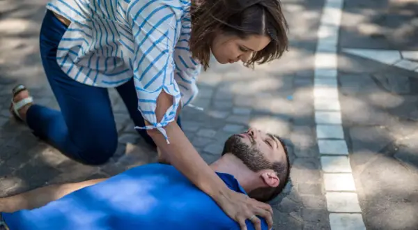 Eine Frau kniet neben einem Mann, der auf dem Boden liegt. Sie prüft seine Atmung, um zu sehen, was passiert bei einem allergischen Schock.