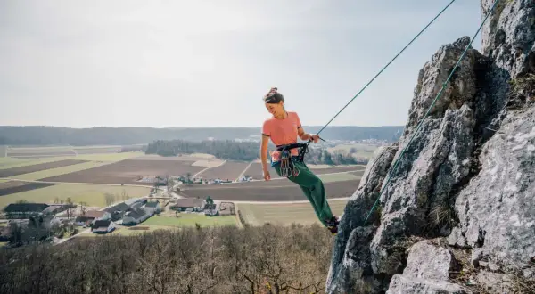 Frau klettert an einem Felsen, Seil ist sicher zum toprope einrichten befestigt. Im Hintergrund Felder und ein Dorf.