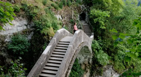Eine Frau genießt die Aussicht auf einer Steinbrücke, während sie in Inzigkofen wandert. Treppen führen zu einer Höhle.