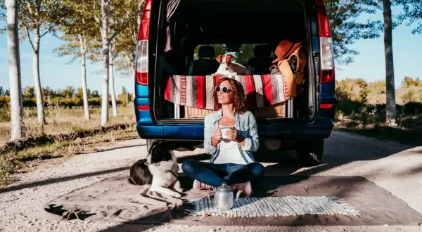 Frau mit Hunden genießt Kaffee vor ihrem Van. Tolle Camping-Hacks für entspannte Ausflüge mit Haustieren.