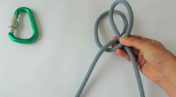 Eine Hand formt einen Knoten aus einem Seil, daneben liegt ein grüner Karabiner. Verschiedene Knoten sind wichtig für Kletterer.