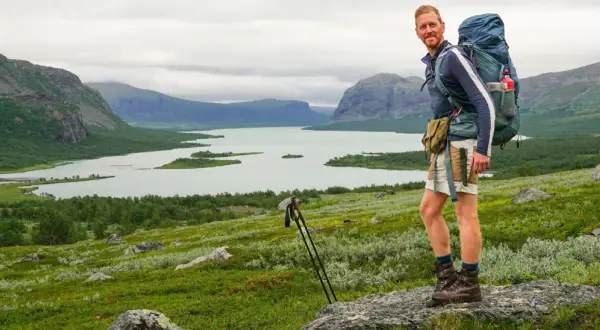 Wanderer mit Rucksack genießt die weite Landschaft im Nationalpark Sarek. Berge, See und grüne Hänge prägen das Bild.