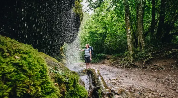 Wanderin unter einem Wasserfall im Schwarzwald. Moosbedeckte Felsen und üppiger Wald säumen den Weg.