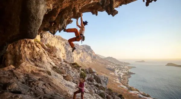 Zwei Frauen beim Klettern an einer Felswand über dem Meer. Die obere Kletterin hängt unter einem Überhang, die untere sichert sie. Verschiedene Kletterarten sind hier zu sehen.