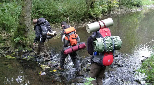 Drei Wanderer durchqueren einen Bach. Ein Mann befestigt seinen Schlafsack am Rucksack.