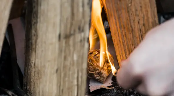 Holzscheite umgeben ein kleines Bündel Zunder, das gerade entzündet wird. Die Flammen tanzen und beginnen, das Holz zu verzehren. Ein Finger ist im Vordergrund zu sehen, der das Feuer macht.