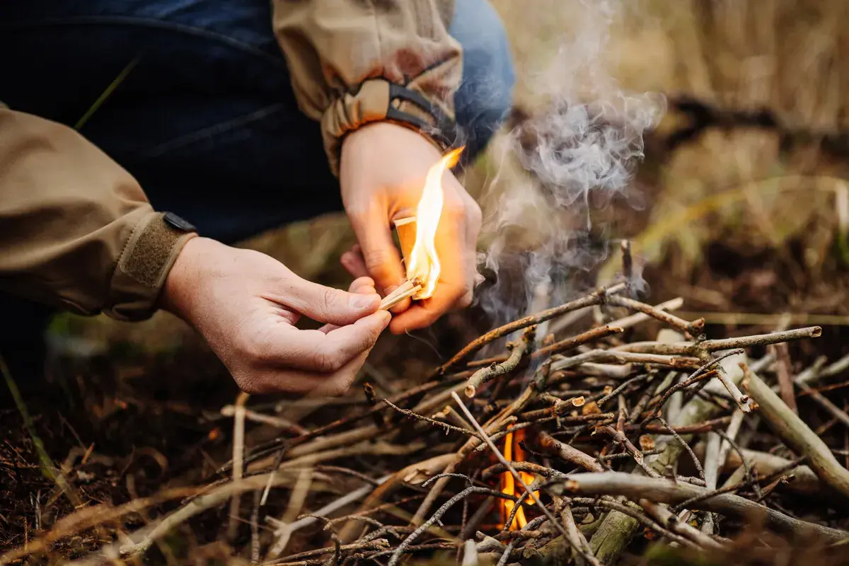 Hände entzünden mit einem Streichholz ein kleines Feuer aus Ästen. Das Lagerfeuer machen beginnt.