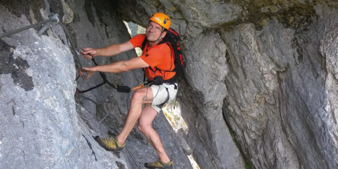Mann klettert gesichert im toten Gebirge. Er trägt Helm, orangefarbenes Shirt und Rucksack.