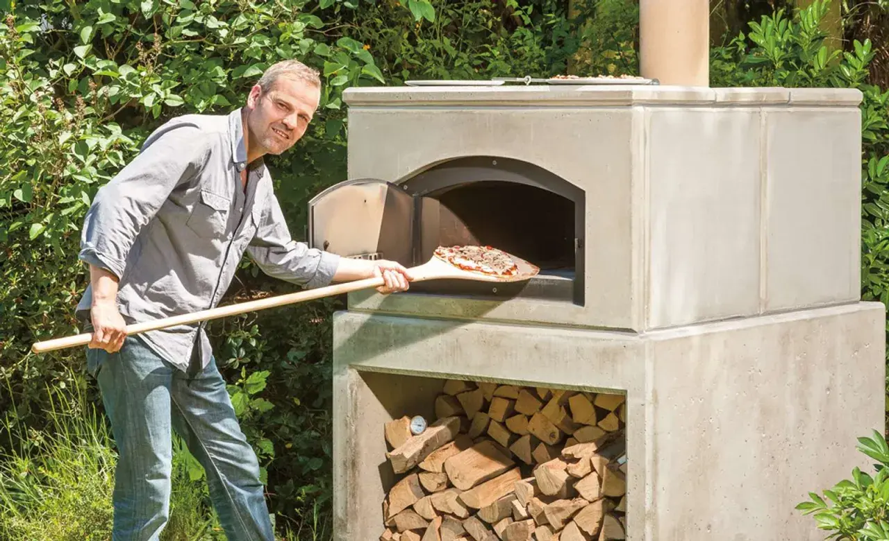 Mann schiebt Pizza in selbstgebauten Outdoor-Ofen. Holz stapelt sich darunter.
