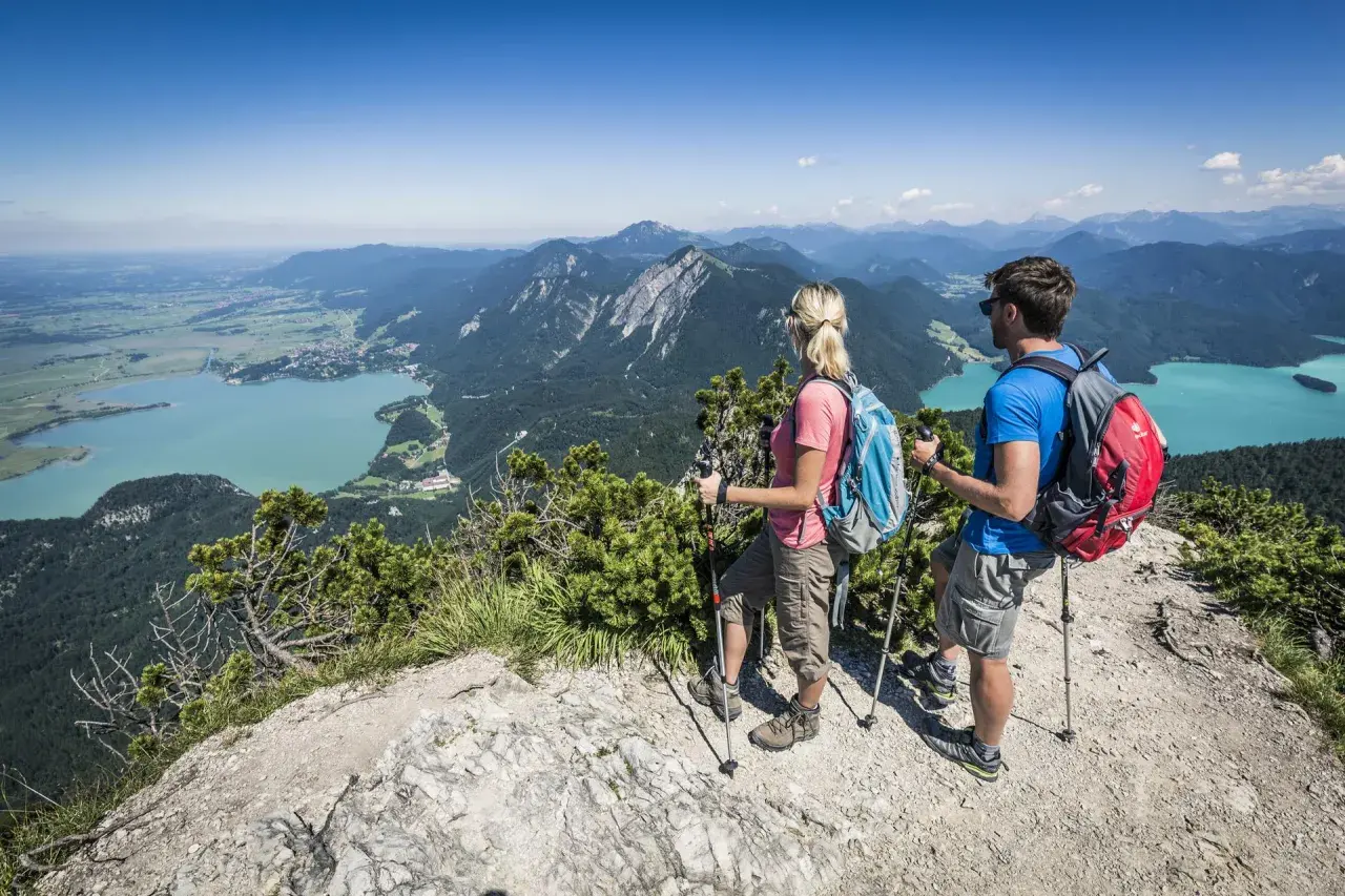 Ein Paar genießt die Aussicht beim Wandern in Mittenwald. Türkisfarbene Seen und grüne Berge erstrecken sich unter ihnen.