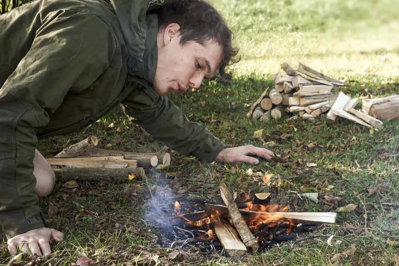 Mann entfacht mit einem Feuerstahl ein Lagerfeuer. Er pustet vorsichtig auf die Flammen, um sie zu nähren.
