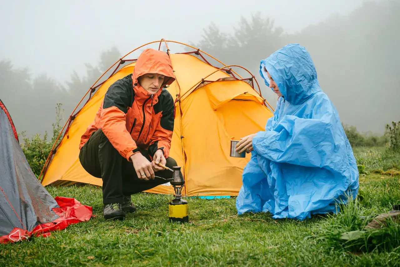 Zwei Personen im Regen bauen ein Zelt auf und bereiten sich ein warmes Getränk zu.