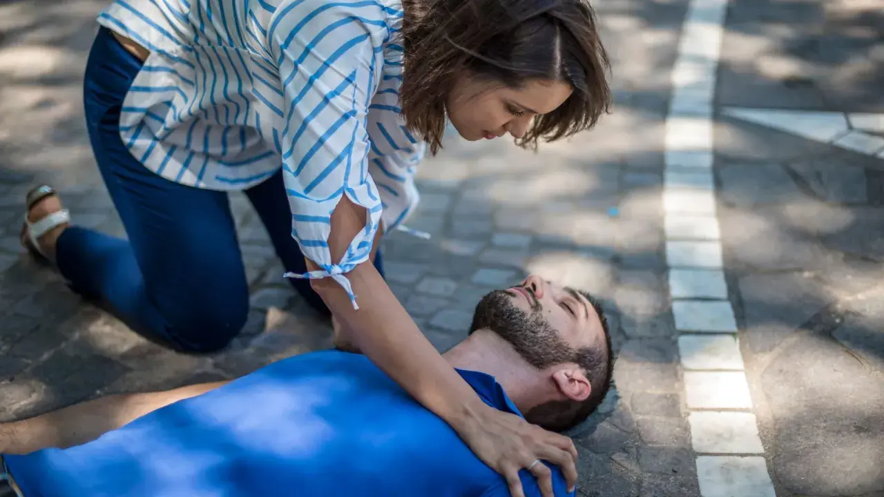Eine Frau kniet neben einem Mann, der auf dem Boden liegt. Sie prüft seine Atmung, um zu sehen, was passiert bei einem allergischen Schock.