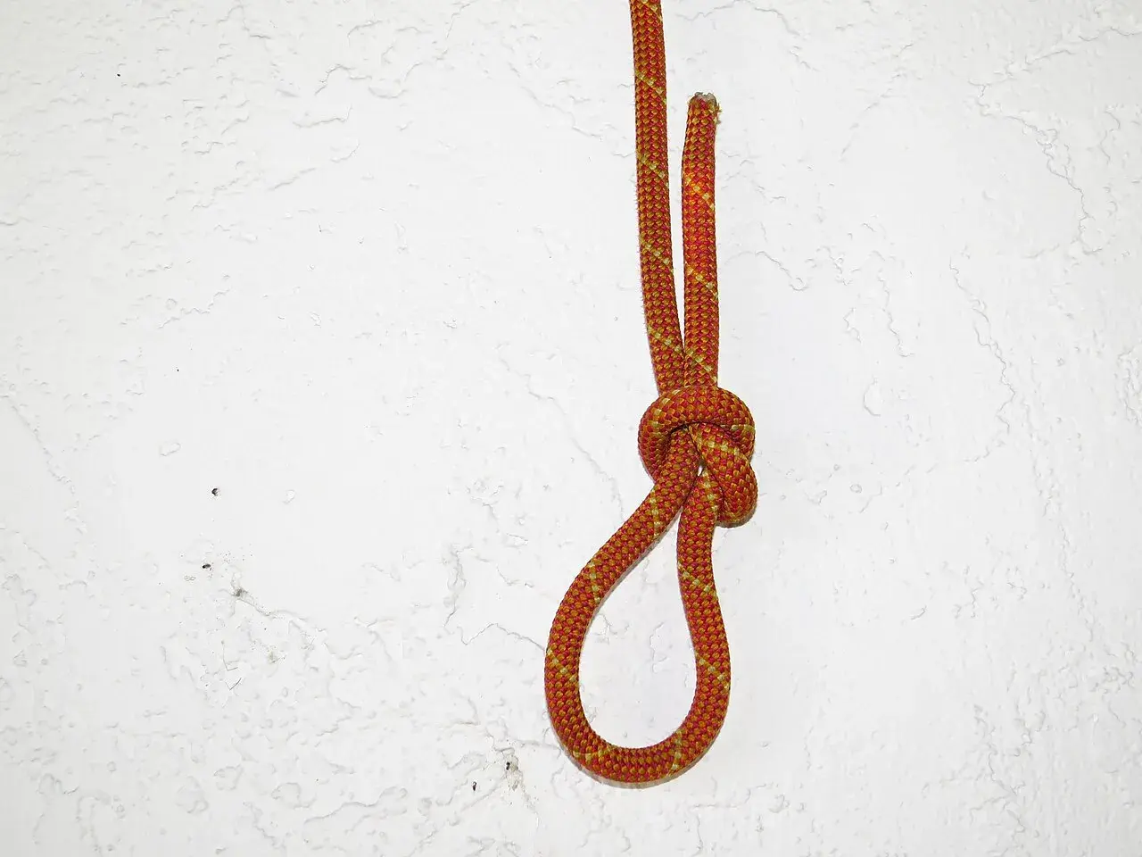 Ein orange-gelbes Seil bildet einen Knoten, der sich zuzieht, vor einer weißen, strukturierten Wand.