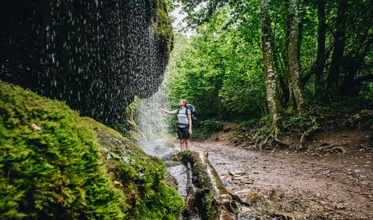 Wanderin unter einem Wasserfall im Schwarzwald. Moosbedeckte Felsen und üppiger Wald säumen den Weg.
