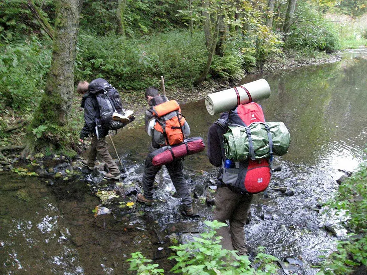 Drei Wanderer durchqueren einen Bach. Ein Mann befestigt seinen Schlafsack am Rucksack.