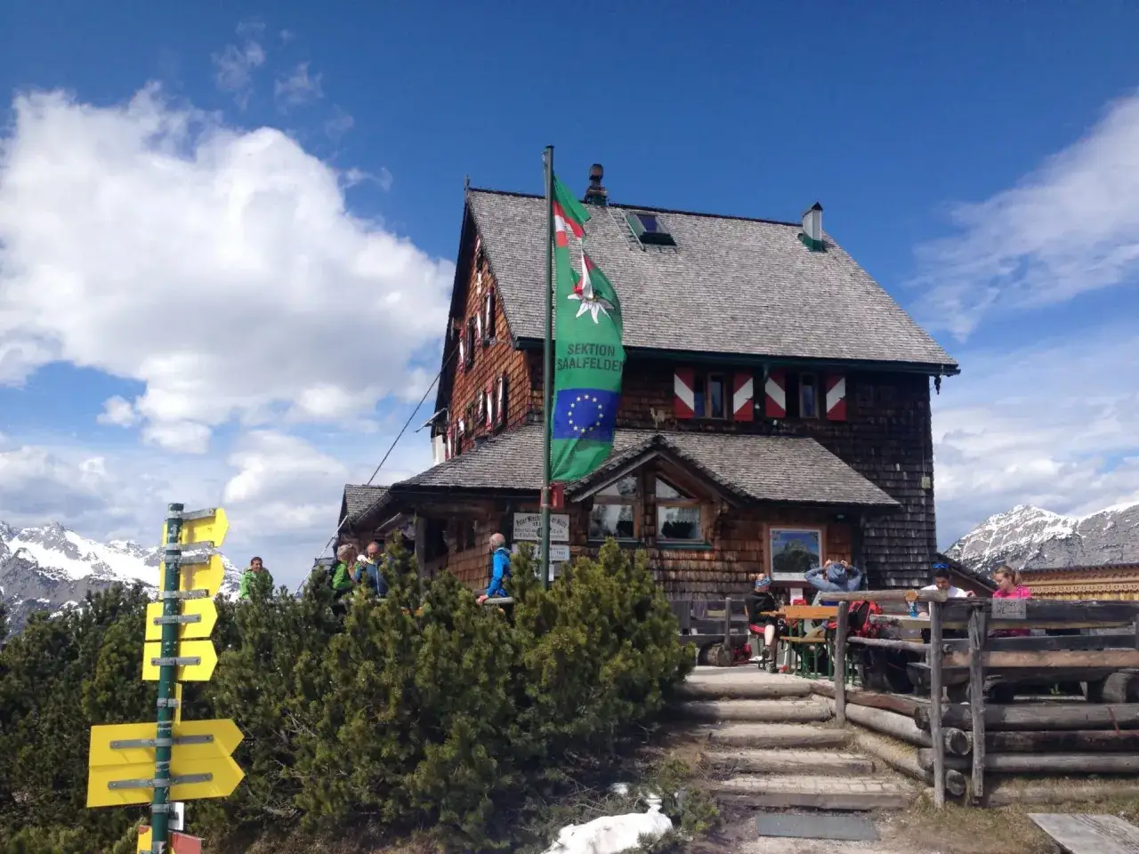 Gemütliche Wanderung mit Einkehr in einer Berghütte. Menschen sitzen draußen, genießen die Aussicht und die Sonne.