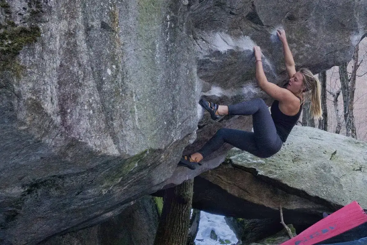 Eine Frau klettert an einem Felsen im Tessin. Sie macht Bouldern und nutzt kleine Griffe, um sich hochzuziehen.