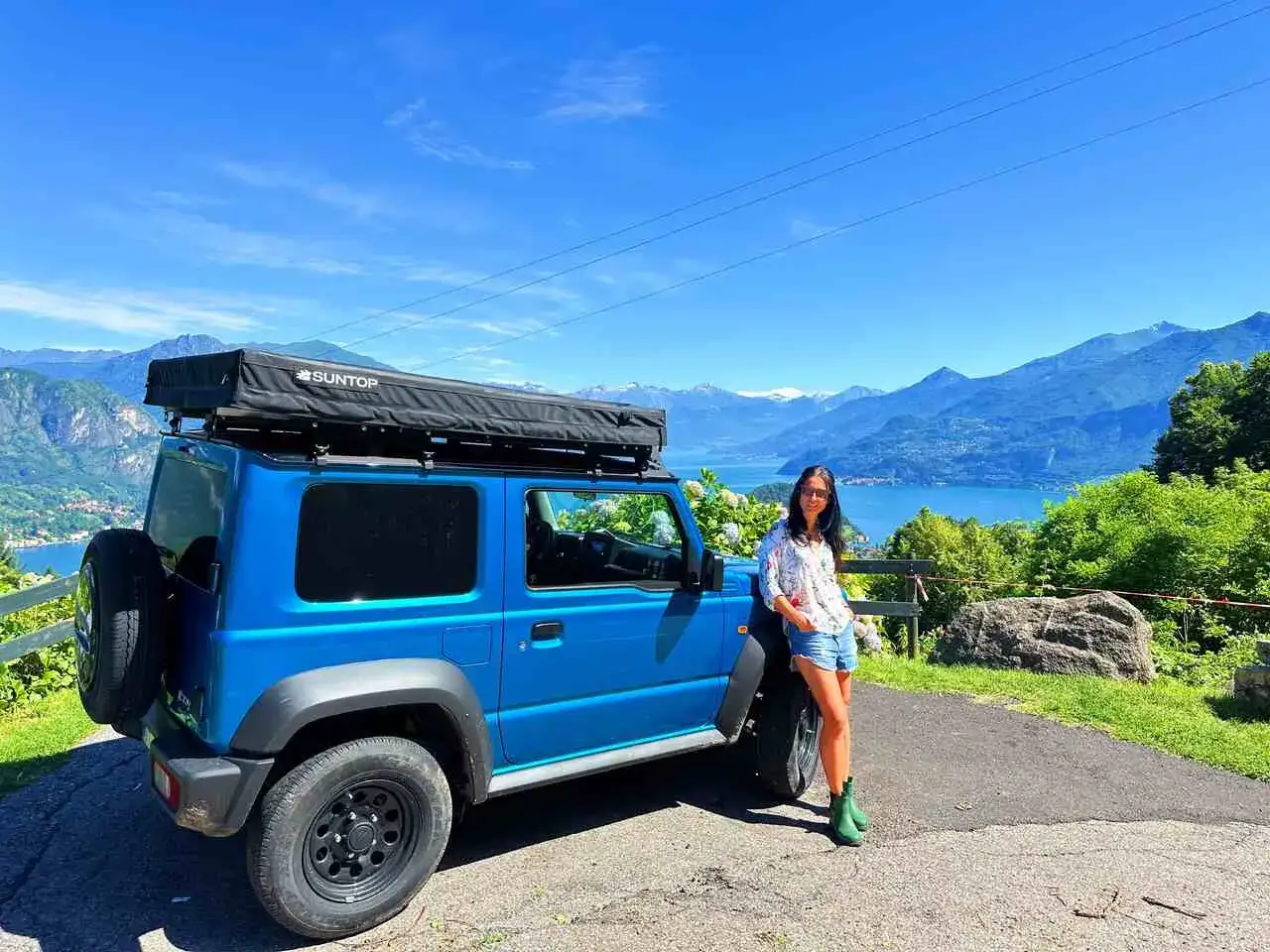 Frau lehnt an blauem Geländewagen mit Dachzelt, Blick auf See und Berge. Hier darf man in Italien im Auto schlafen, ein Traum!