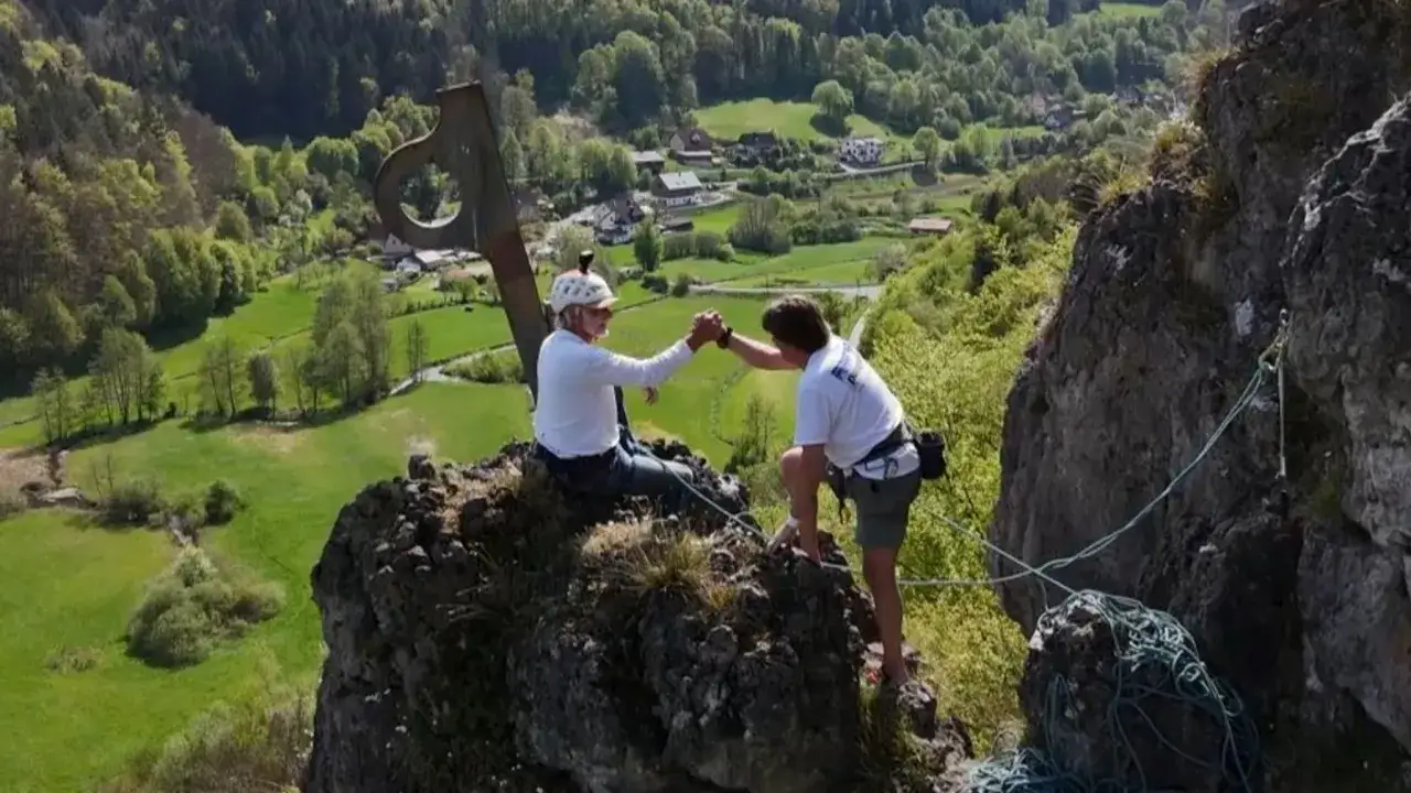 Zwei Kletterer geben sich nach erfolgreichem rotpunkt klettern am Gipfel die Hand. Im Hintergrund ein malerisches Tal.