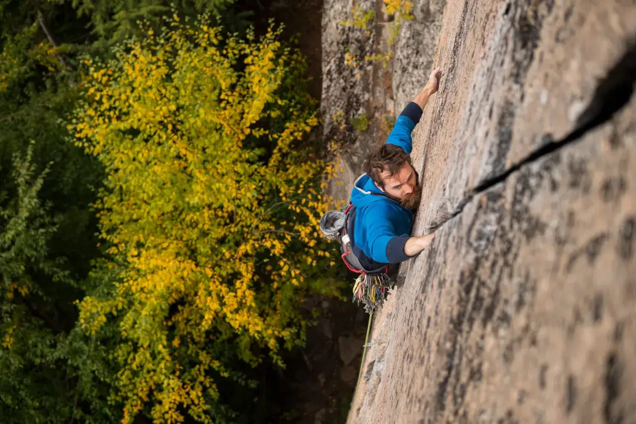 Ein Mann klettert eine Felswand hoch, im Hintergrund leuchtet Herbstlaub. Er nutzt eine breite Spalte für sein offwidth climbing.
