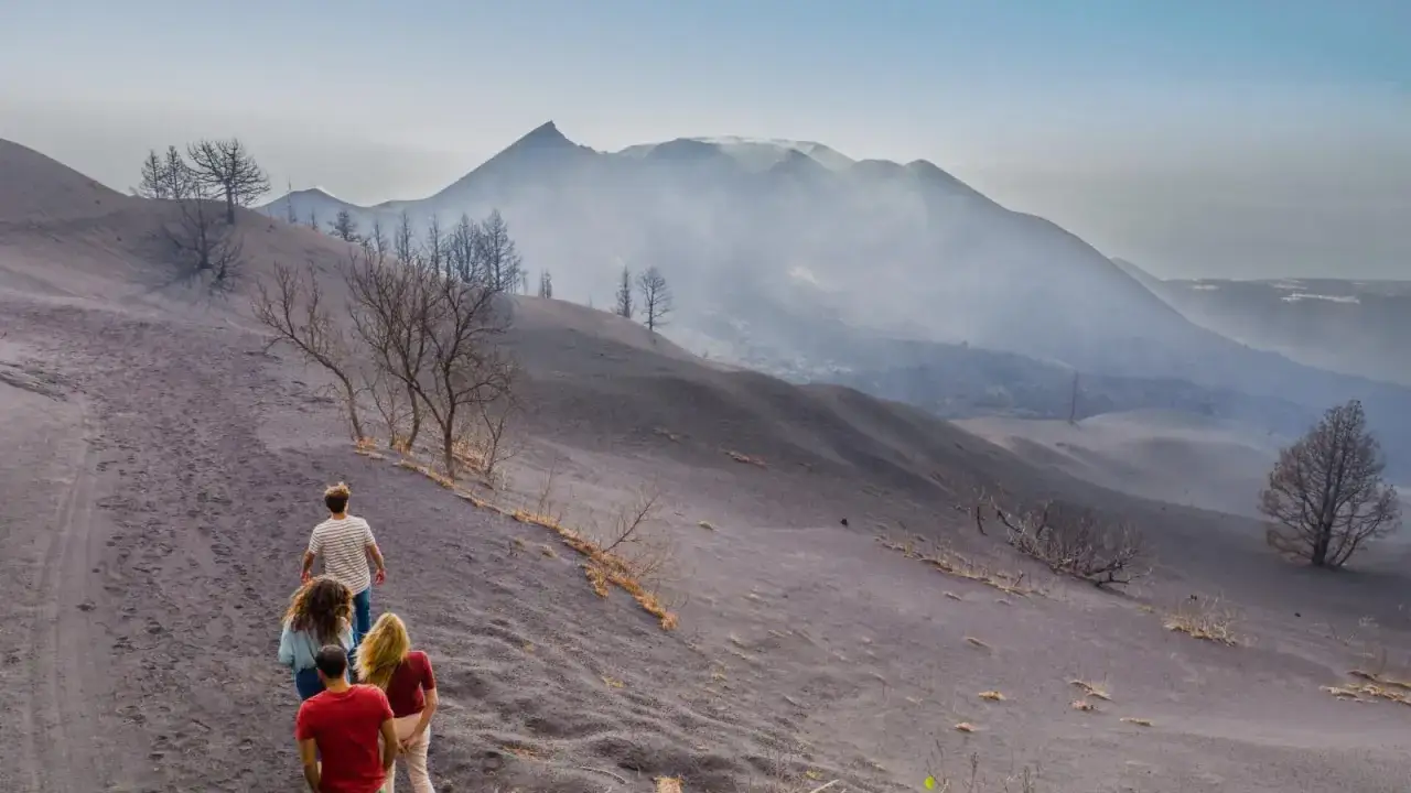 Vier Personen wandern auf einem Pfad durch eine aschebedeckte Landschaft auf La Palma. Im Hintergrund raucht ein Vulkan.