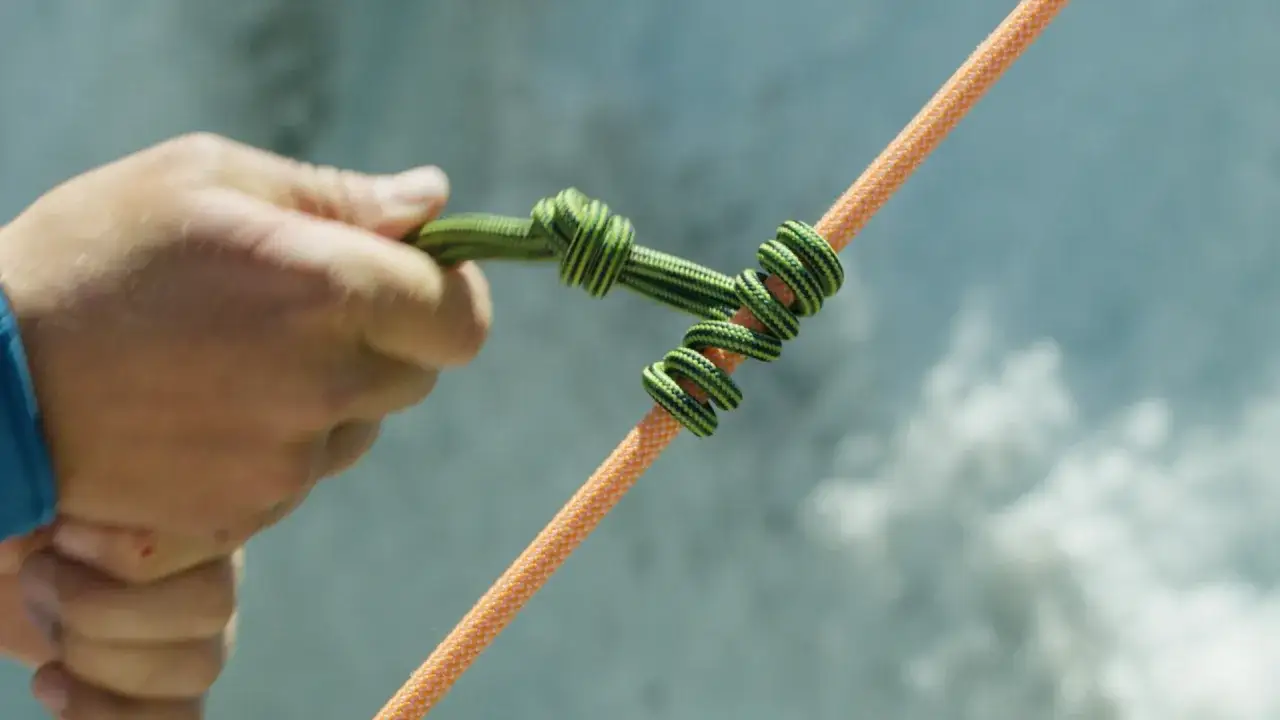 Eine Hand bindet einen HMS Knoten auf ein oranges Seil.