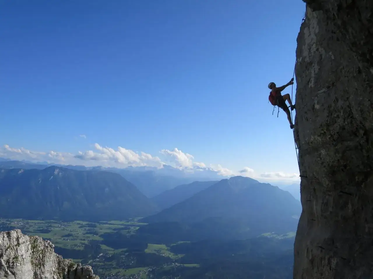 Kletterer am toten Gebirge, mit Blick auf ein grünes Tal und blaue Berge unter strahlend blauem Himmel.