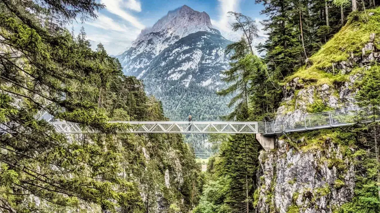 Ein Wanderer genießt die Aussicht von einer Hängebrücke über einer Schlucht, umgeben von Bäumen, mit einem majestätischen Berg im Hintergrund. Ein tolles Erlebnis beim Wandern in Mittenwald.