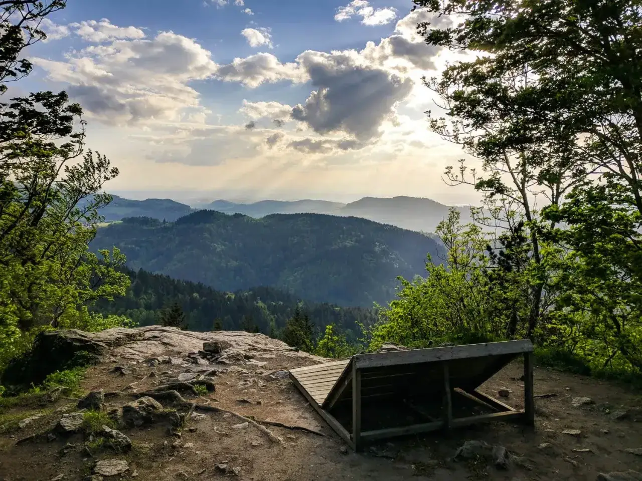 Sonnenstrahlen brechen durch Wolken über einem grünen Tal. Ein Holzpodest lädt zum Verweilen ein. Perfekt für eine Auszeit beim Wandern Freiburg.