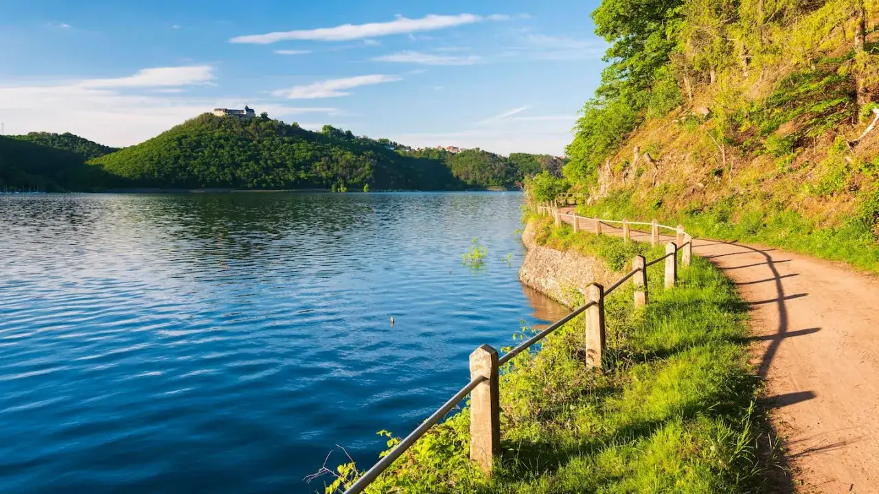 Ein Weg lädt zum wandern am Wasser ein. Grüne Hügel mit einer Burgruine im Hintergrund und ein blauer See prägen die Szenerie.