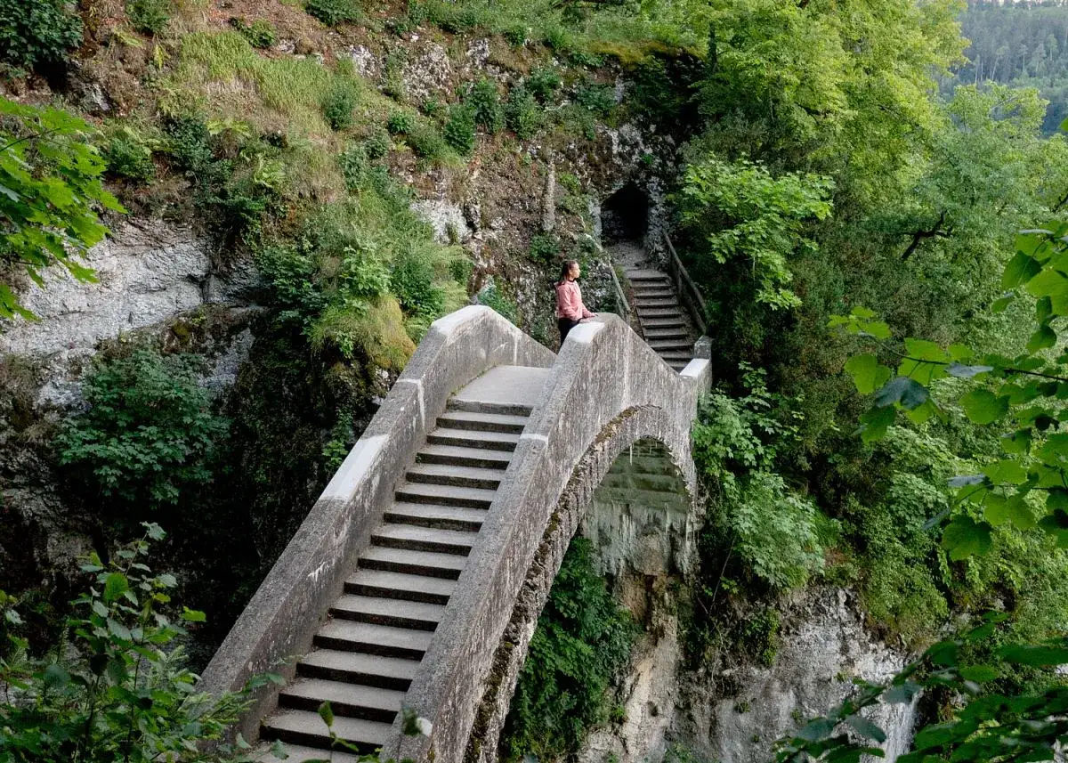 Eine Frau genießt die Aussicht auf einer Steinbrücke, während sie in Inzigkofen wandert. Treppen führen zu einer Höhle im grünen Berghang.