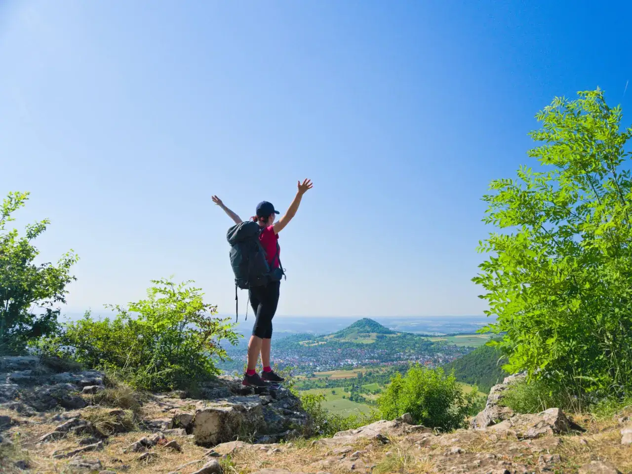 Wanderin mit Rucksack auf einem Felsen, Arme erhoben, genießt die Aussicht auf das Tal. Die HW1 Etappen sind geschafft!