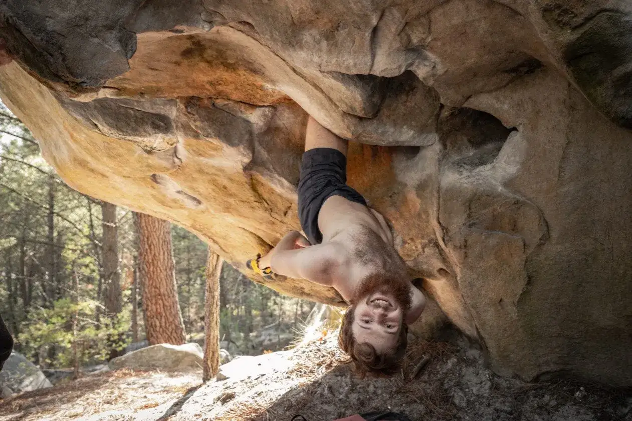 Ein Mann hängt kopfüber an einem Felsen und lächelt. Er klettert eine Route, die zu den anspruchsvolleren Boulder Grades gehört.