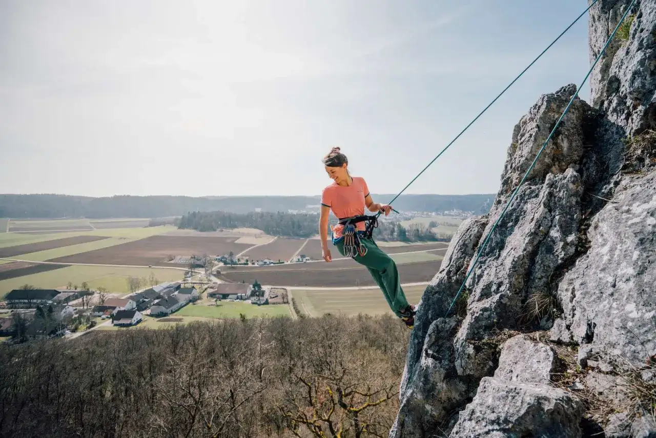 Frau seilt sich nur mit Seil an einem Felsen ab, mit Blick auf ein malerisches Dorf und Felder.