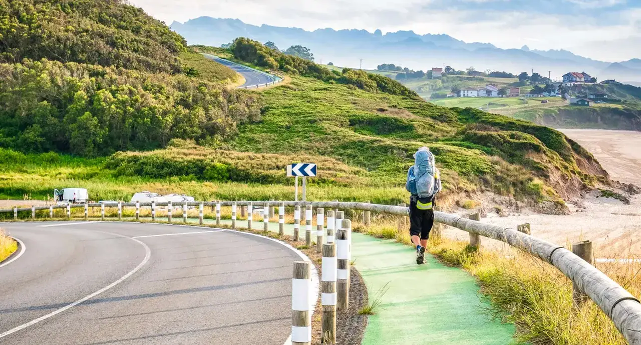 Wanderer mit Rucksack auf einem Küstenweg, der Alternativen zum Jakobsweg erkundet. Grüne Hügel, Straße und Meer im Hintergrund.