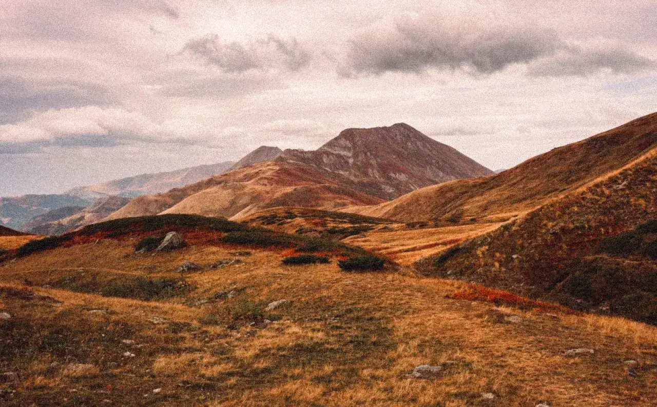 Herbstliche Berglandschaft mit einem high scardus trail. Goldbraune Gräser und rote Büsche bedecken die Hänge unter einem dramatischen Himmel.