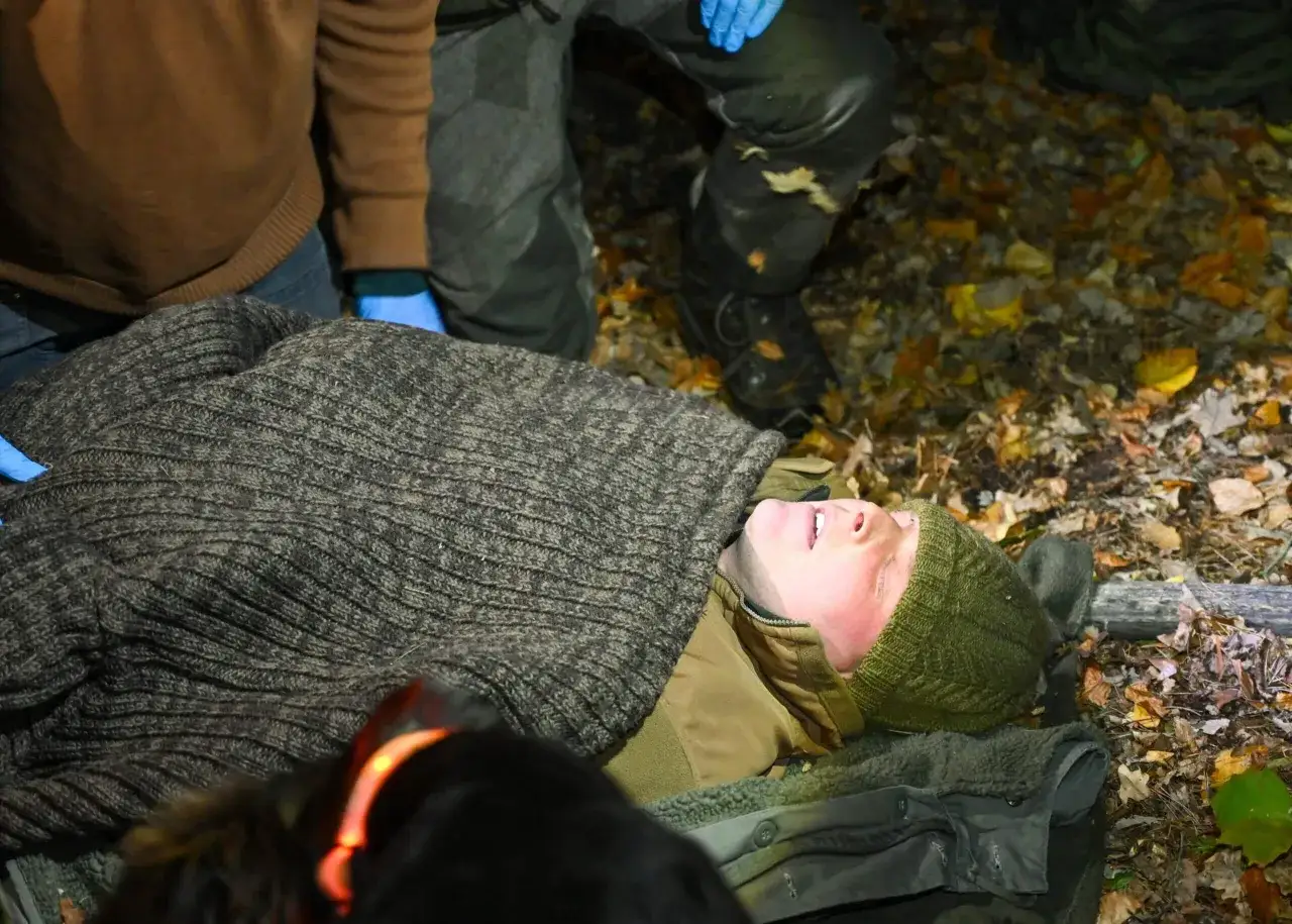 Person liegt auf dem Boden, zugedeckt mit einer Decke. Ein Hund mit leuchtendem Halsband ist im Vordergrund. Die Szene könnte auf eine schlangenbiss wunde hindeuten.