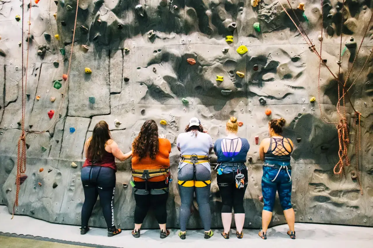 F&uuml;nf Frauen in Klettergurten und bequemer Kleidung stehen vor einer Kletterwand. Sie sind bereit zu sehen, was man zum Klettern anzieht.