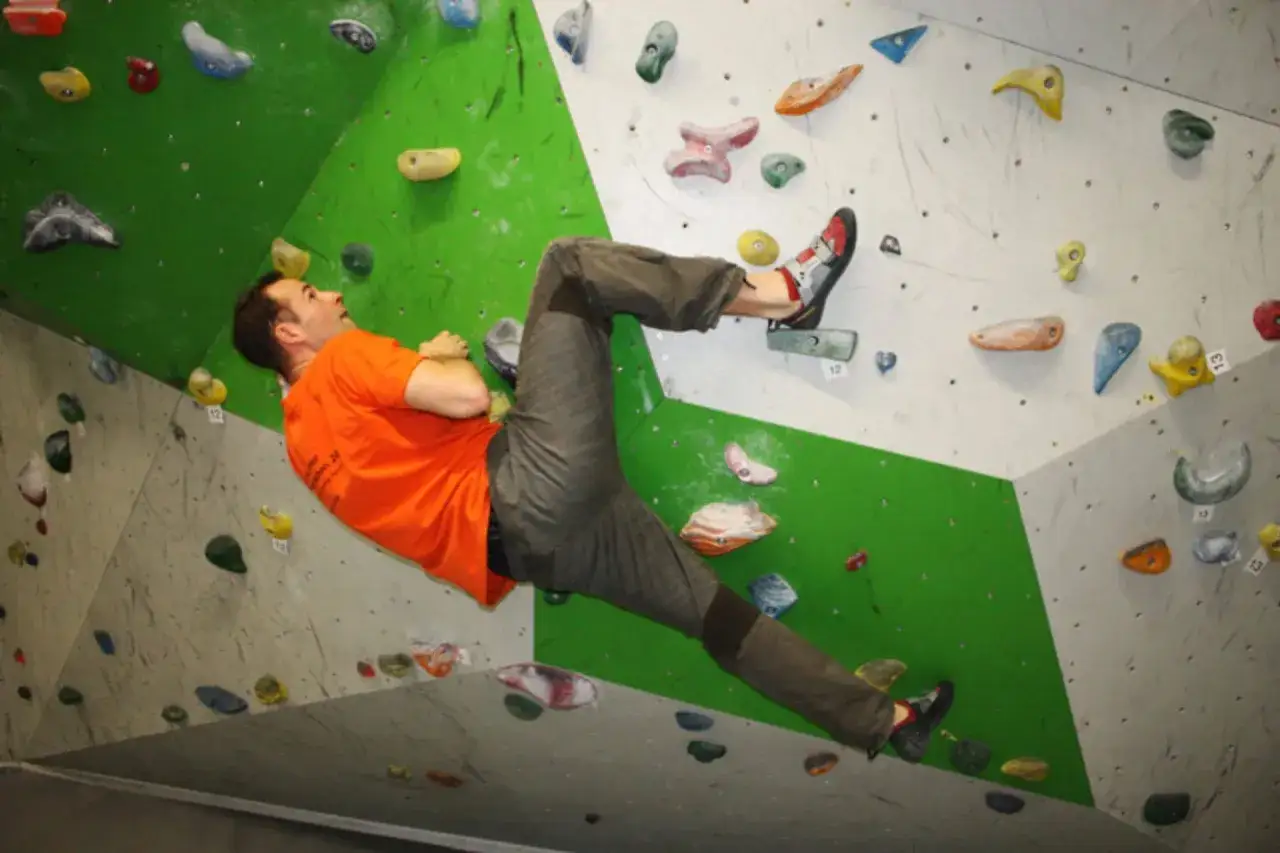 Mann klettert an einer Boulderwand, Schwierigkeitsgrad 4. Er nutzt bunte Griffe, um sich vorwärts zu bewegen.