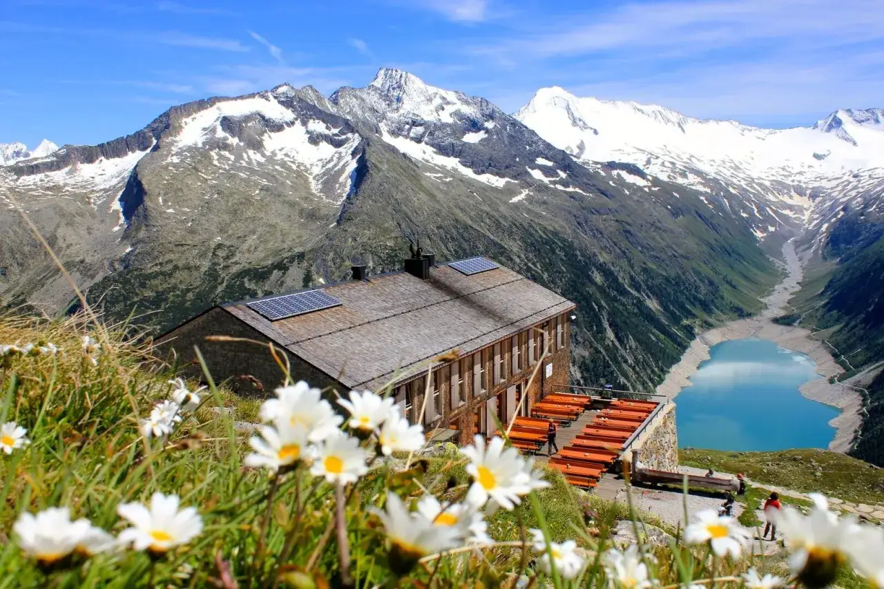 Alpenhütte mit Solaranlagen, davor blühende Wiesen und im Hintergrund ein Stausee. Ein perfekter Ort für die Peter Habeler Runde.