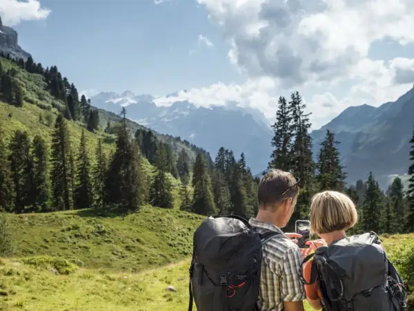 Ein Paar plant seine **Wanderung** in den Alpen. Sie schauen auf ihr Handy, um die Route zu prüfen, umgeben von grünen Hängen und schneebedeckten Gipfeln.