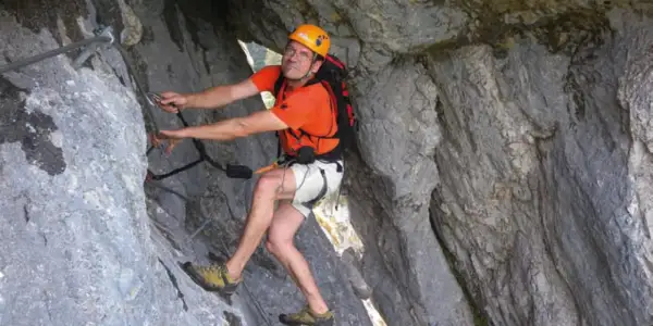 Mann klettert gesichert im toten Gebirge. Er trägt Helm, orangefarbenes Shirt und Rucksack.