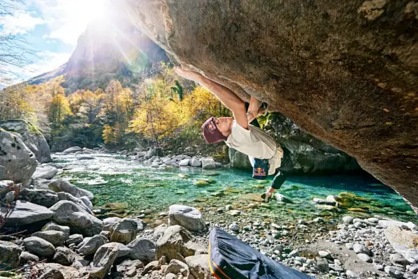 Ein Kletterer meistert eine anspruchsvolle Route beim **Tessin Bouldern** über einem kristallklaren Fluss. Herbstlaub und Berge im Hintergrund.