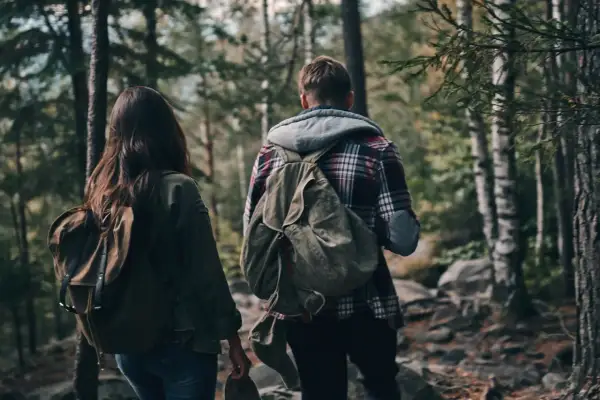 Zwei Personen mit Rucksäcken wandern durch einen Wald. Ein Mann und eine Frau, die sich auf ein Abenteuer vorbereiten, das über einfaches Hiking hinausgeht und eher dem Trekking ähnelt.