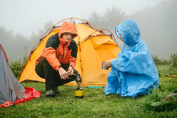 Zwei Personen im Regen bauen ein Zelt auf und bereiten sich ein warmes Getränk zu.